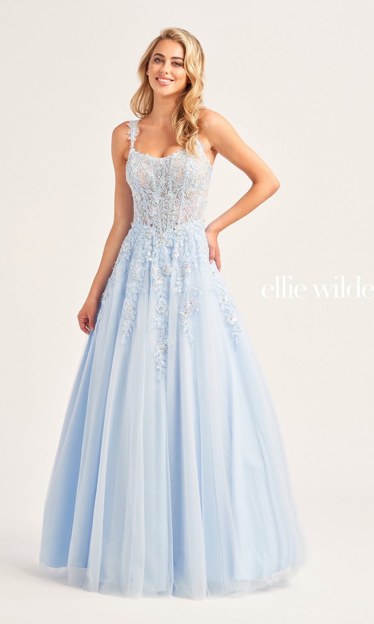 A woman in the Ellie Wilde A-Line Long Designer Ball Gown EW35123—a light blue, floor-length dress with a lace bodice and tulle skirt—stands against a plain background.